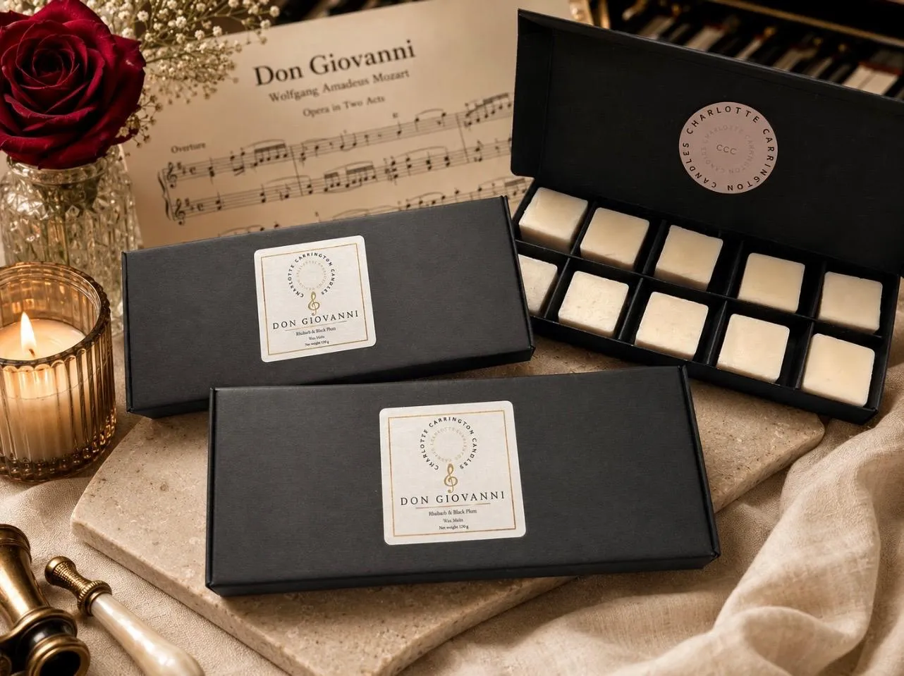 A selection of wax melts presentation boxes sitting on cloth with a candle, a rose in a vase and a sheet of music